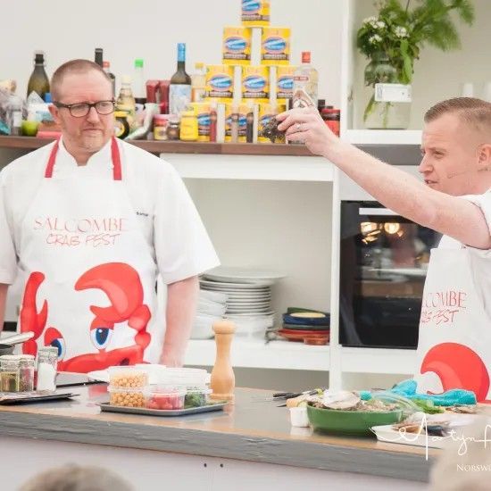 Two chefs in white aprons with lobster logos cooking in a kitchen. One holds up a small glass.