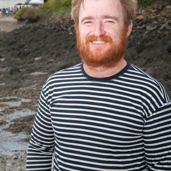Man with red beard, wearing striped shirt, smiling outdoors near water.
