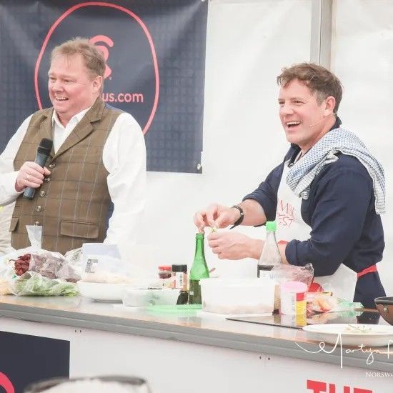 Two men at a cooking demonstration. One speaks into a mic, the other cooks, laughing. White apron, food on counter.