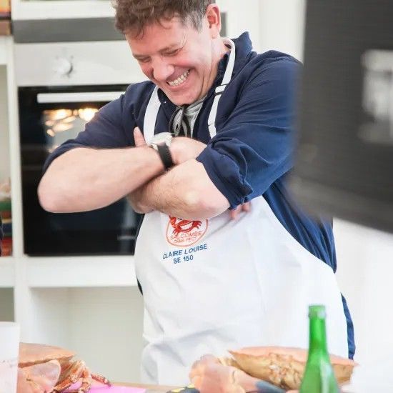 Man in apron laughing, arms crossed, in a kitchen with an oven in the background.