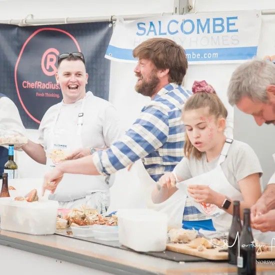 People preparing food at an outdoor event. Man laughs, others work at a counter. Banner reads 
