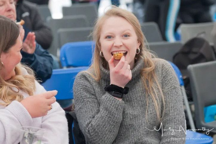 Woman smiling, eating food at outdoor event, wearing gray sweater. Others in background.