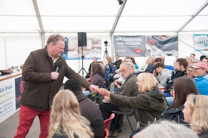 Man in brown jacket speaking into a microphone, reaching to shake hands with a woman in a seated audience, outdoors in tent.