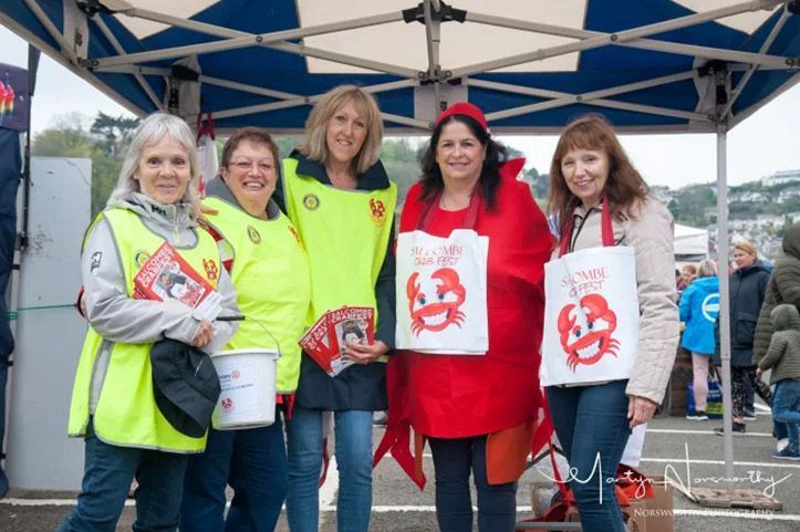 Five women in front of a tent, fundraising. One wears a crab costume. They hold bags with a crab logo, smiling.