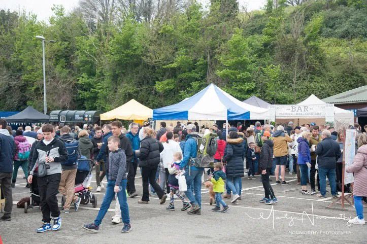 Crowd at outdoor market with tents. People walk around, looking at stalls. Trees in the background.