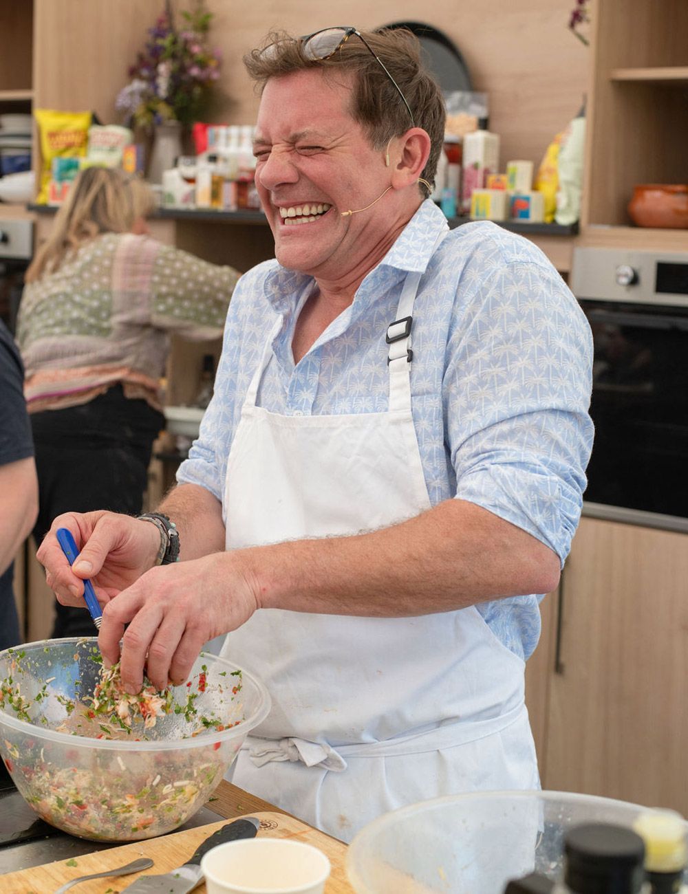 Man in apron laughing, stirring food in a bowl in a kitchen.