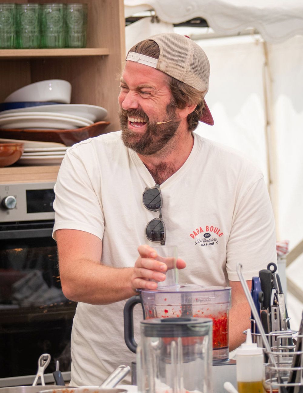 Man in a baseball cap laughs while using a food processor in a kitchen setting.