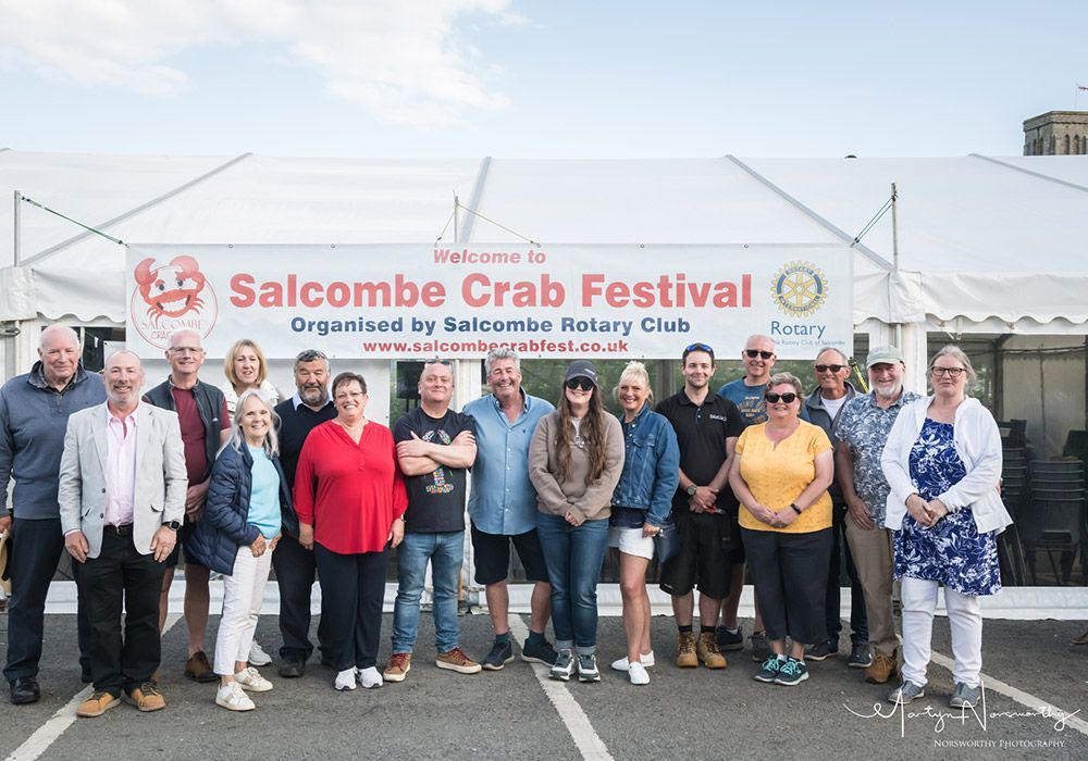 Group poses in front of a Salcombe Crab Festival banner on a sunny day.