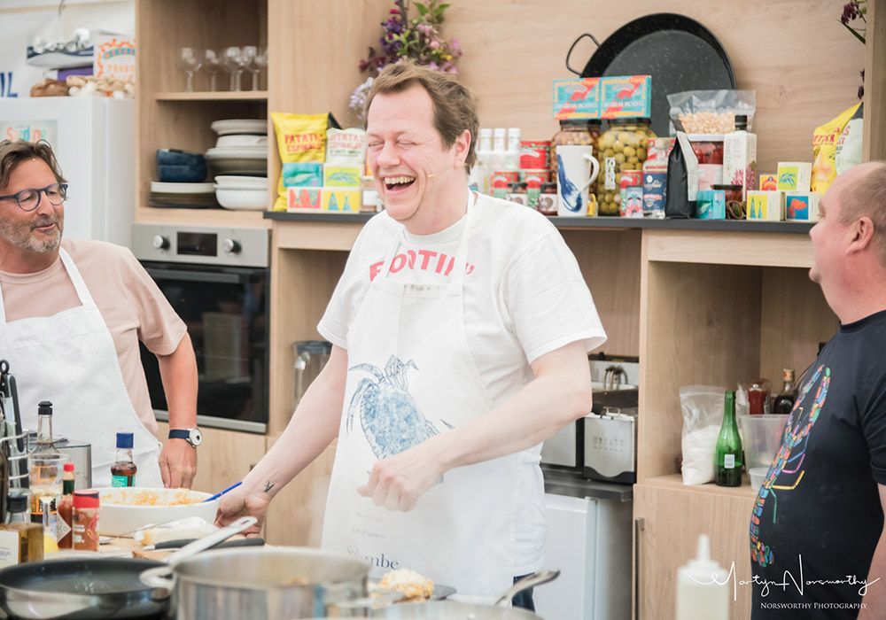 Three men in a kitchen; one laughs while cooking.