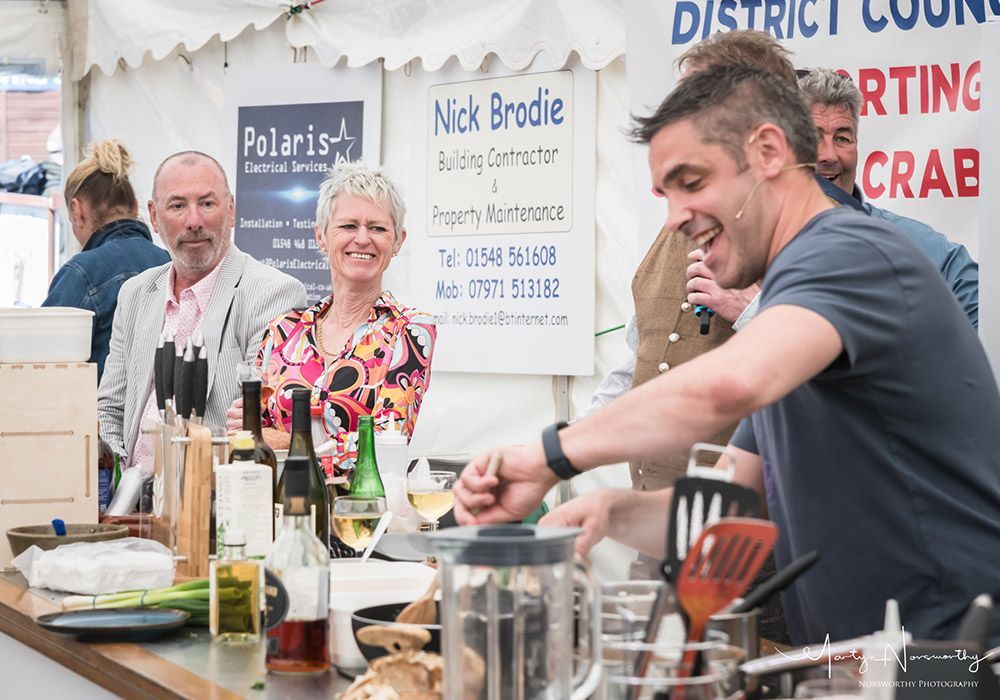 Chef cooking, smiling, at outdoor event. Audience watches. White tent.