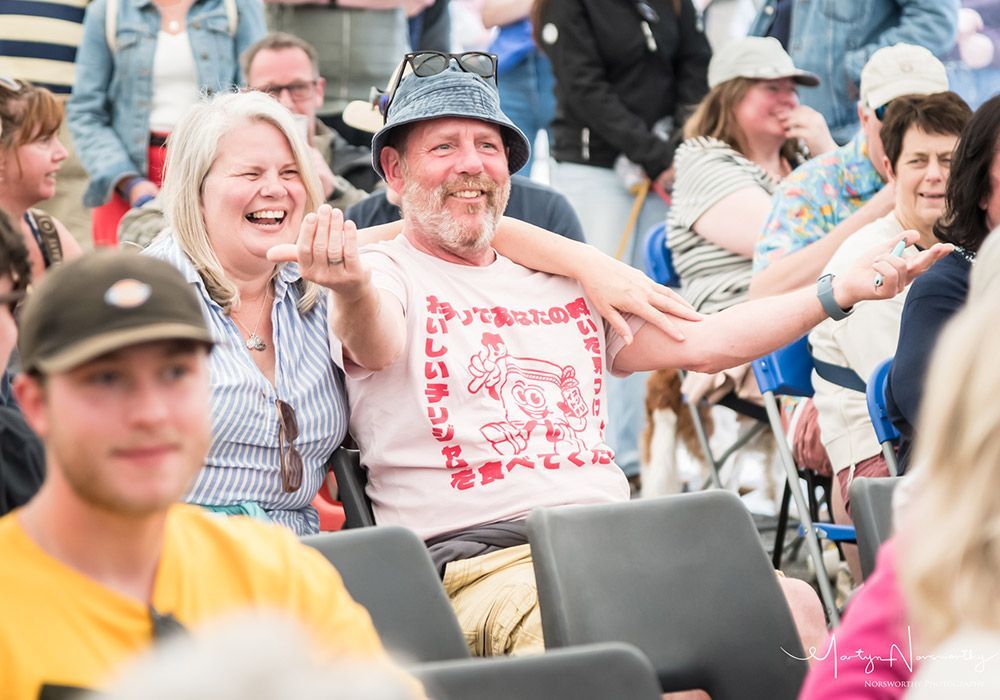 Smiling couple at an outdoor event, man in bucket hat and pink shirt with arms outstretched, woman with arm around him.