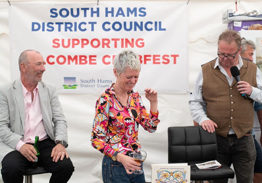 Three people at a Combe Curfest event, one holding a microphone, others seated. Banner in background.