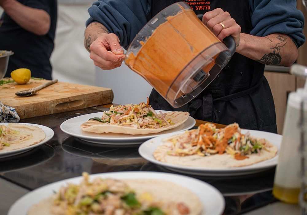 A chef pours sauce from a food processor onto wraps filled with food on plates.