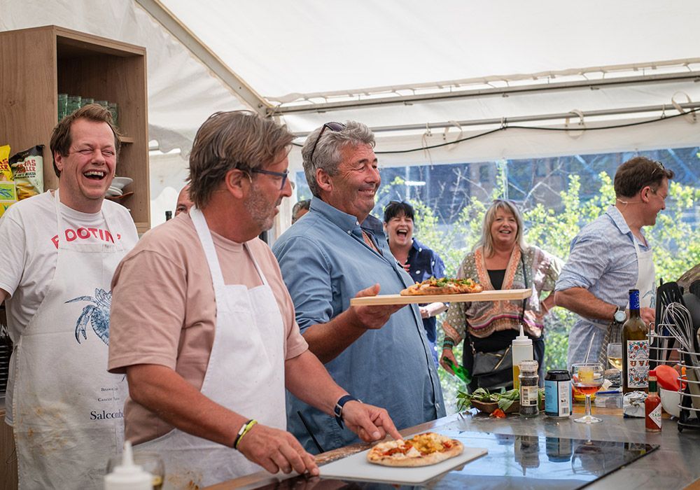People in a kitchen tent laugh and hold pizzas.