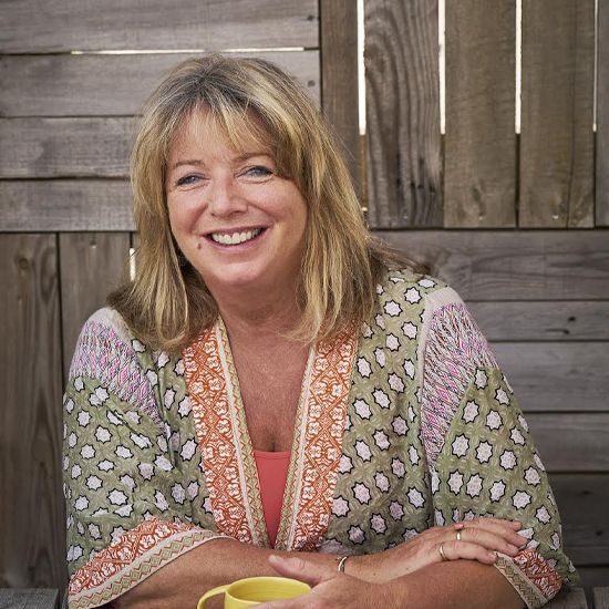 Woman smiling, wearing a patterned top, seated outdoors, wooden fence background, holding a yellow mug.