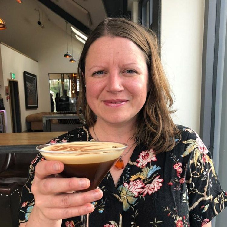 Woman holding a cocktail in a bar; she smiles, and wears a floral top.