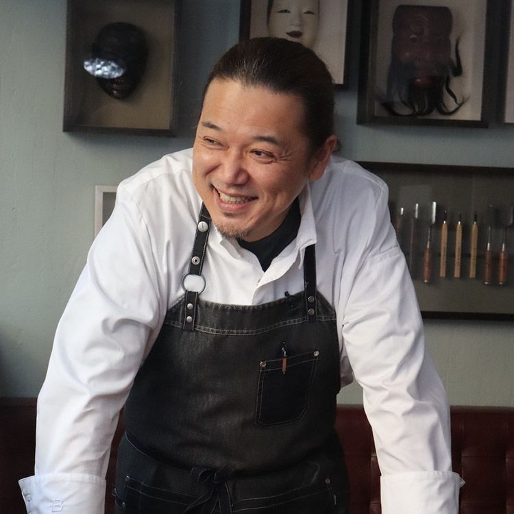 Man in apron smiling, leaning against a surface, with masks and tools on the wall behind him.
