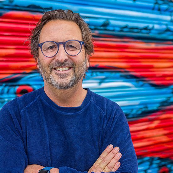 Man wearing glasses smiles, standing in front of colorful mural, blue shirt.