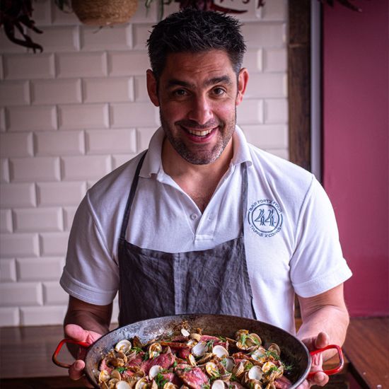 Chef holding a pan of food, smiling. Wearing white shirt and grey apron. Interior setting.