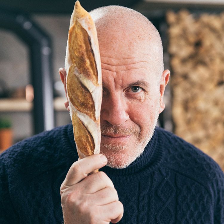 Bald man holding a baguette in front of his face, wearing a dark blue sweater.
