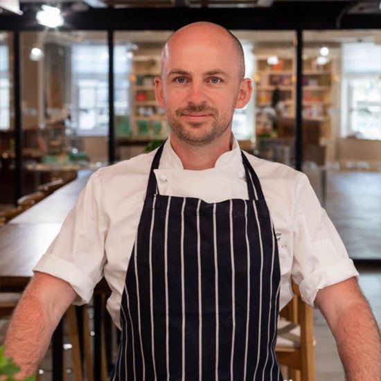 Chef with bald head wearing a white shirt and striped apron stands in a restaurant.