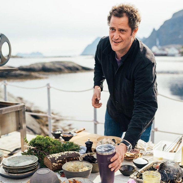 Man reaching for a purple cup at an outdoor table with food, overlooking water and mountains.