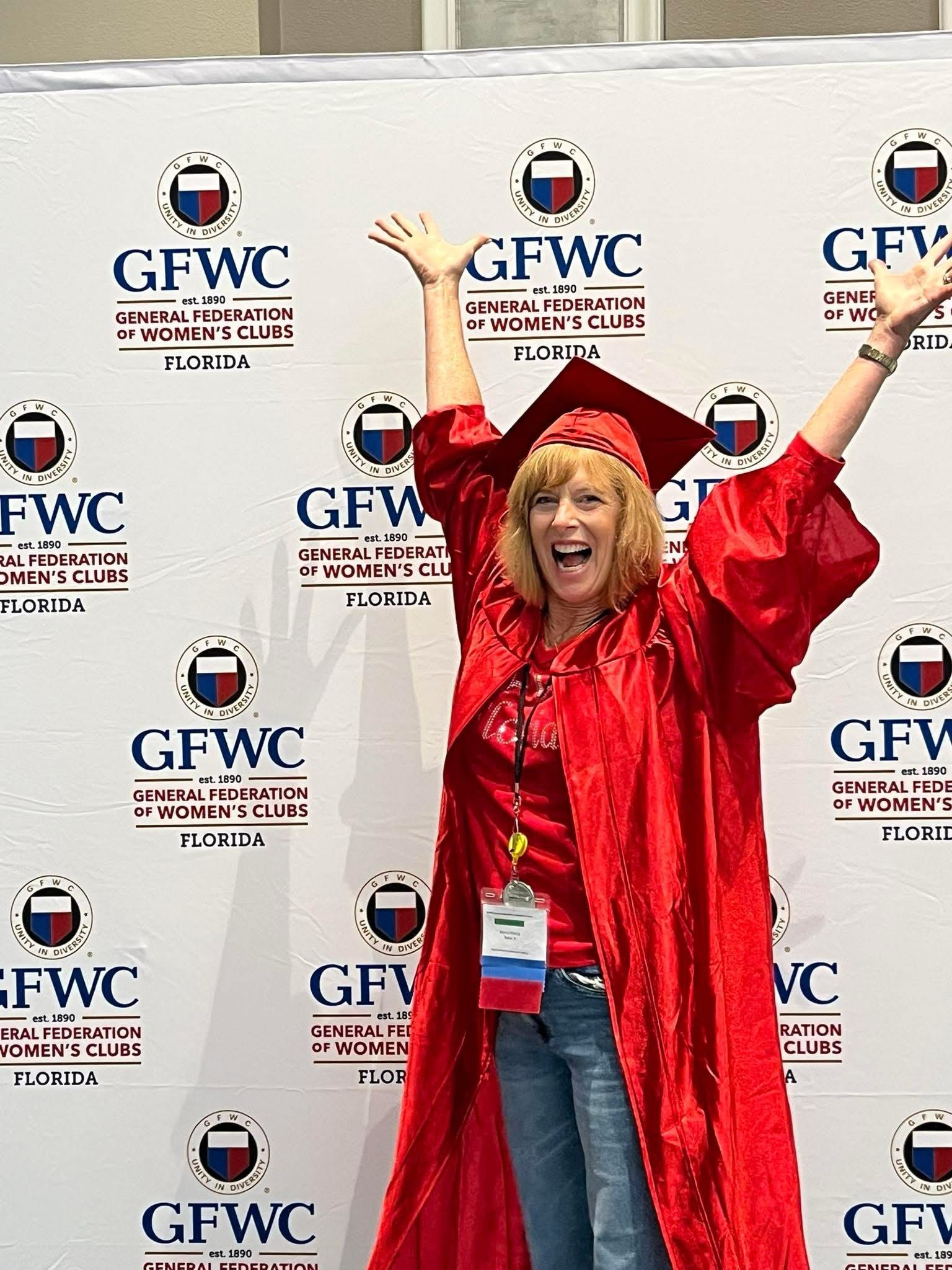 Woman in red graduation gown and cap, arms raised, smiling at a GFWC event backdrop.