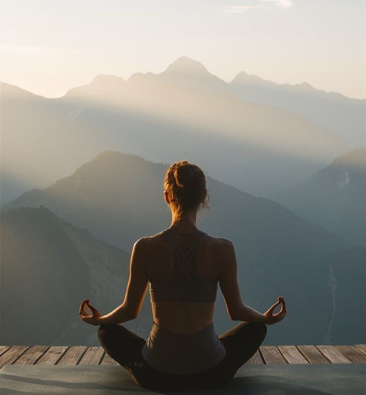 A woman is sitting in a lotus position on a yoga mat with mountains in the background.
