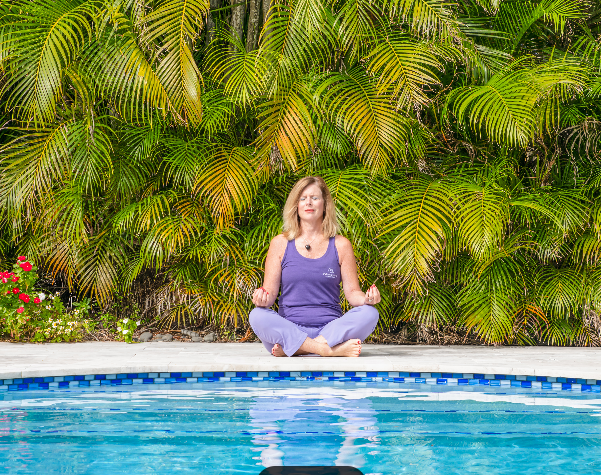 Person sitting cross-legged by a pool in front of lush green tropical plants.