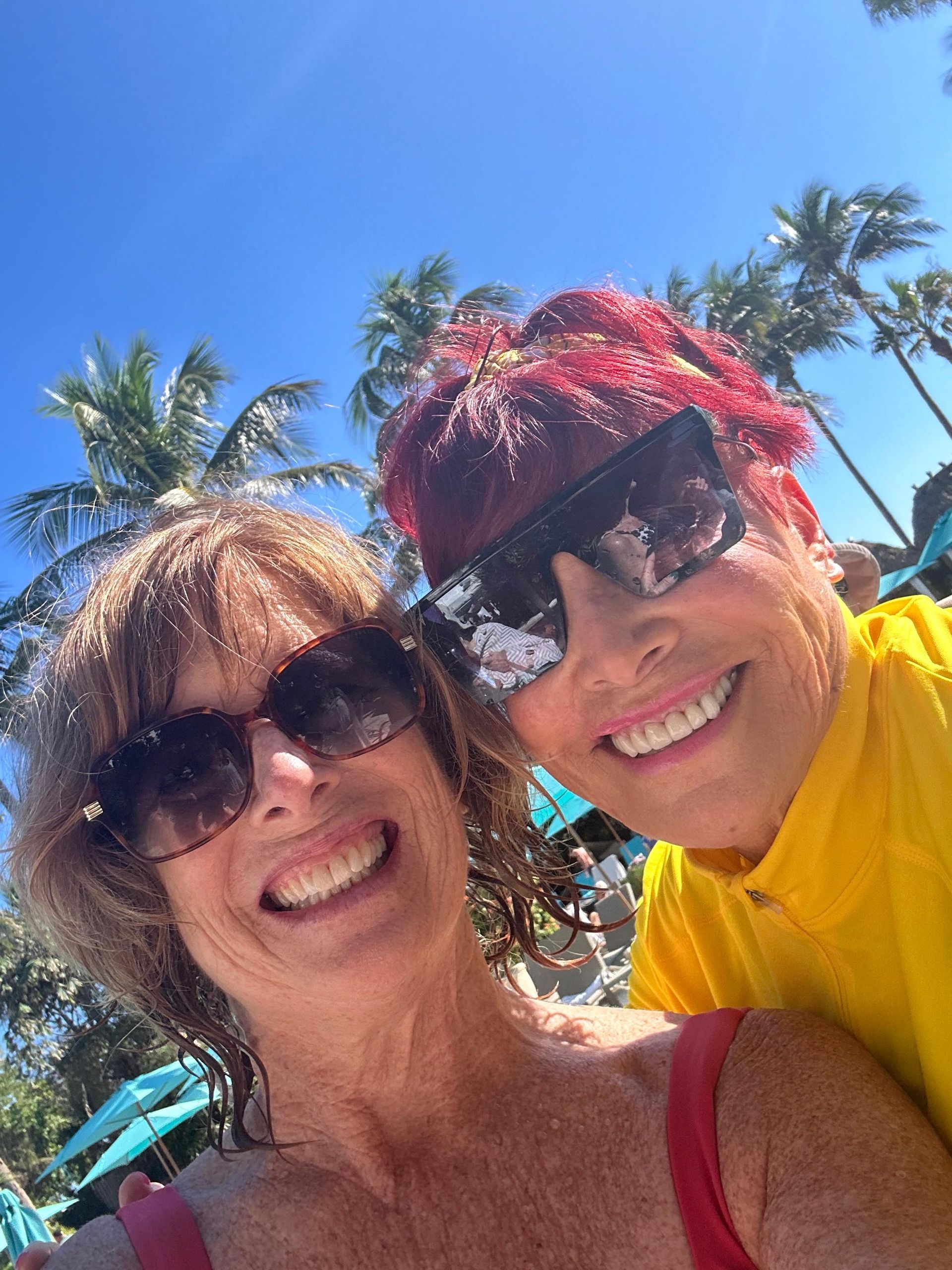 Two women smile for a selfie in sunny outdoor setting with palm trees. One with red hair and yellow shirt.
