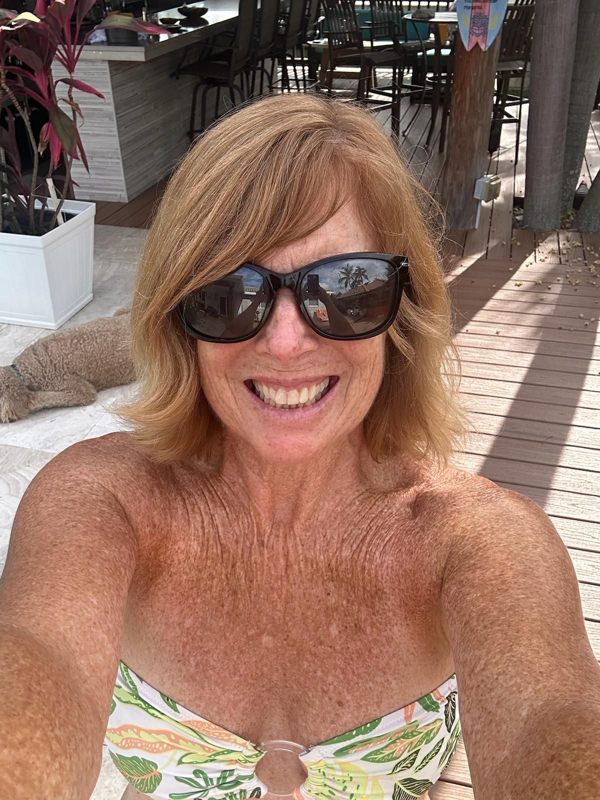 Woman with sunglasses smiles, in a tropical setting, wearing a floral swimsuit.