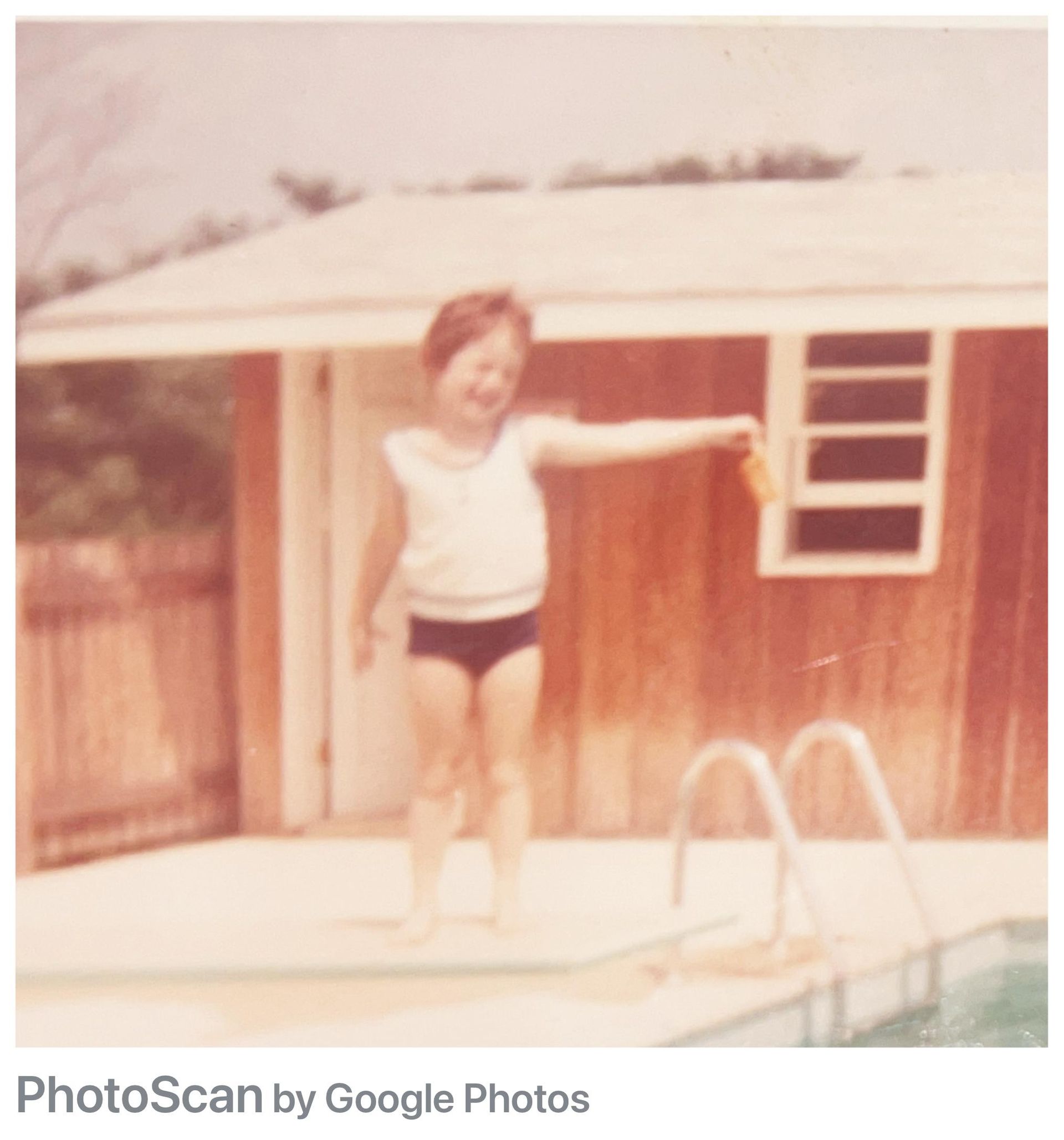 Child in swimsuit by a pool, smiles and reaches, with a small building in the background.
