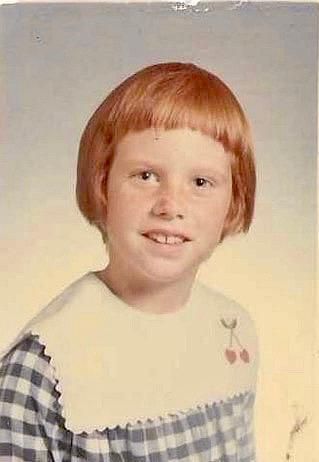 A young girl with red hair and a bowl cut smiles in a school photo, wearing a gingham dress with a white collar.