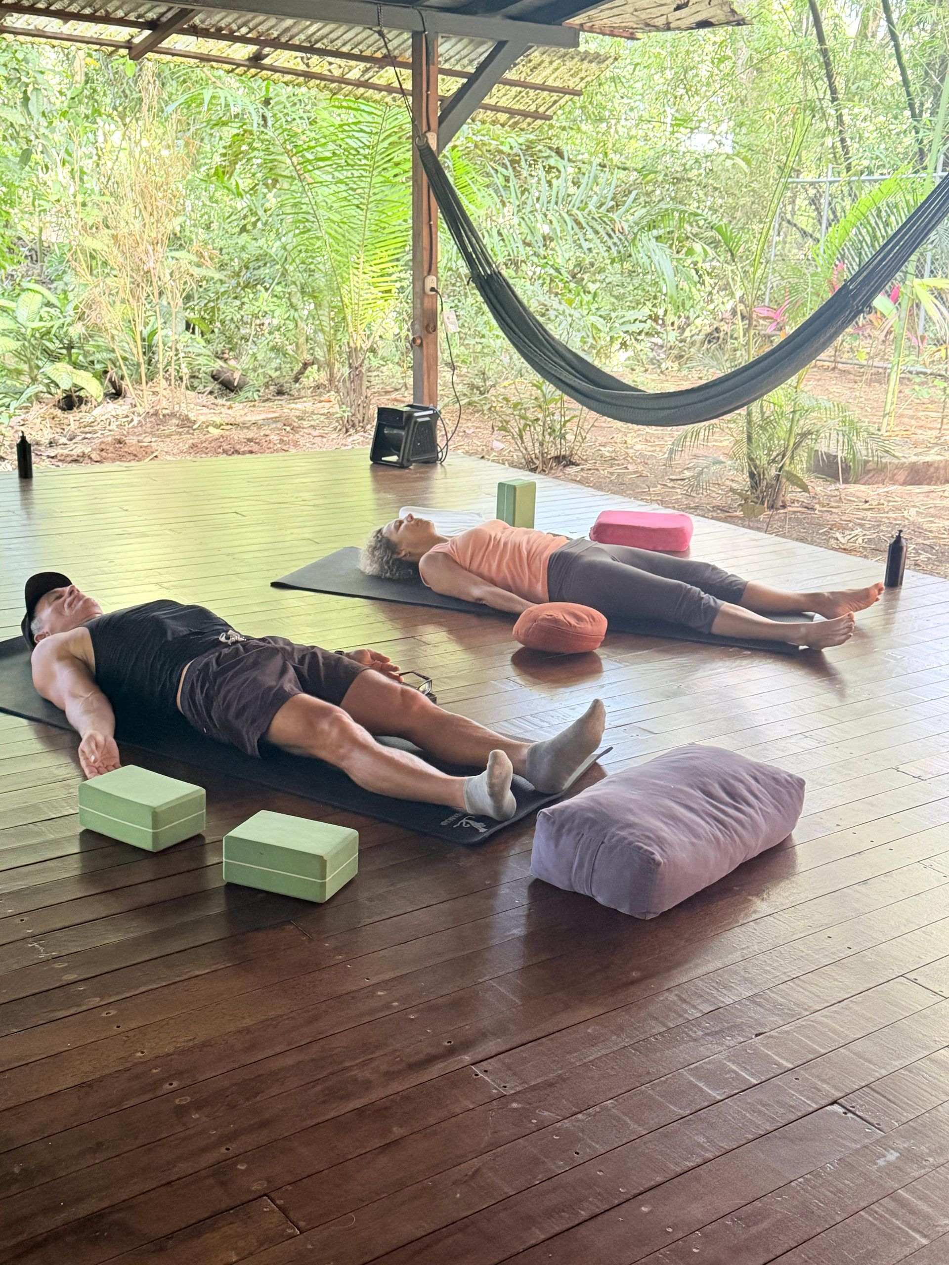 Two people lying on yoga mats in a wooden outdoor pavilion with a hammock nearby