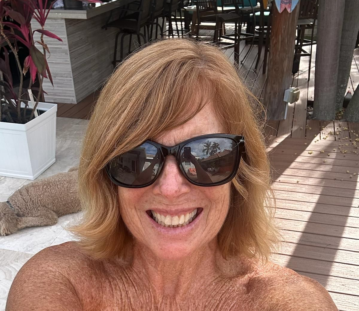 Smiling person in sunglasses on a wooden deck with outdoor seating and potted plants behind them