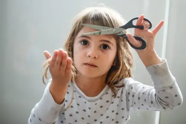 Girl holding scissors near her bangs, looking concerned; some cut hair in hand.