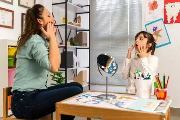 Two women at a desk in a bright room, reacting in surprise while looking at each other and a small mirror.