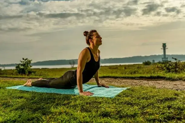 Woman in yoga pose on mat in grassy field by water.
