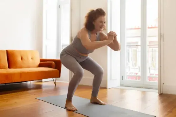 Woman doing a squat on a yoga mat in her living room. She is wearing gray workout clothes.