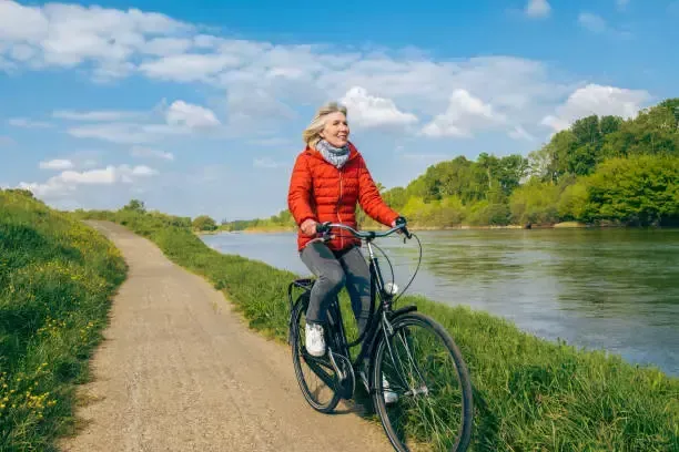 Woman biking on a path next to a river, smiling. She wears red jacket and gray pants on a sunny day.