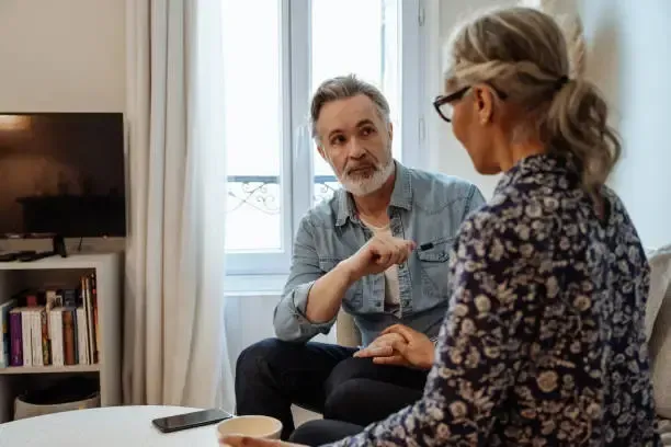 Two people talking on a couch in a bright living room, with a TV and bookshelf in the background