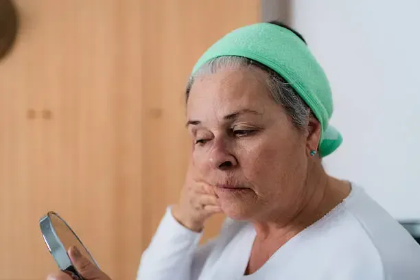 Older woman with grey hair wearing a green headband, looking into a small mirror, touching her face.