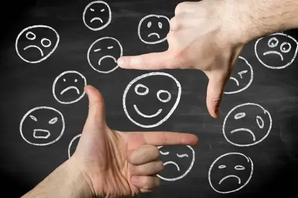 Hands framing a happy face amidst a background of sad faces on a chalkboard.