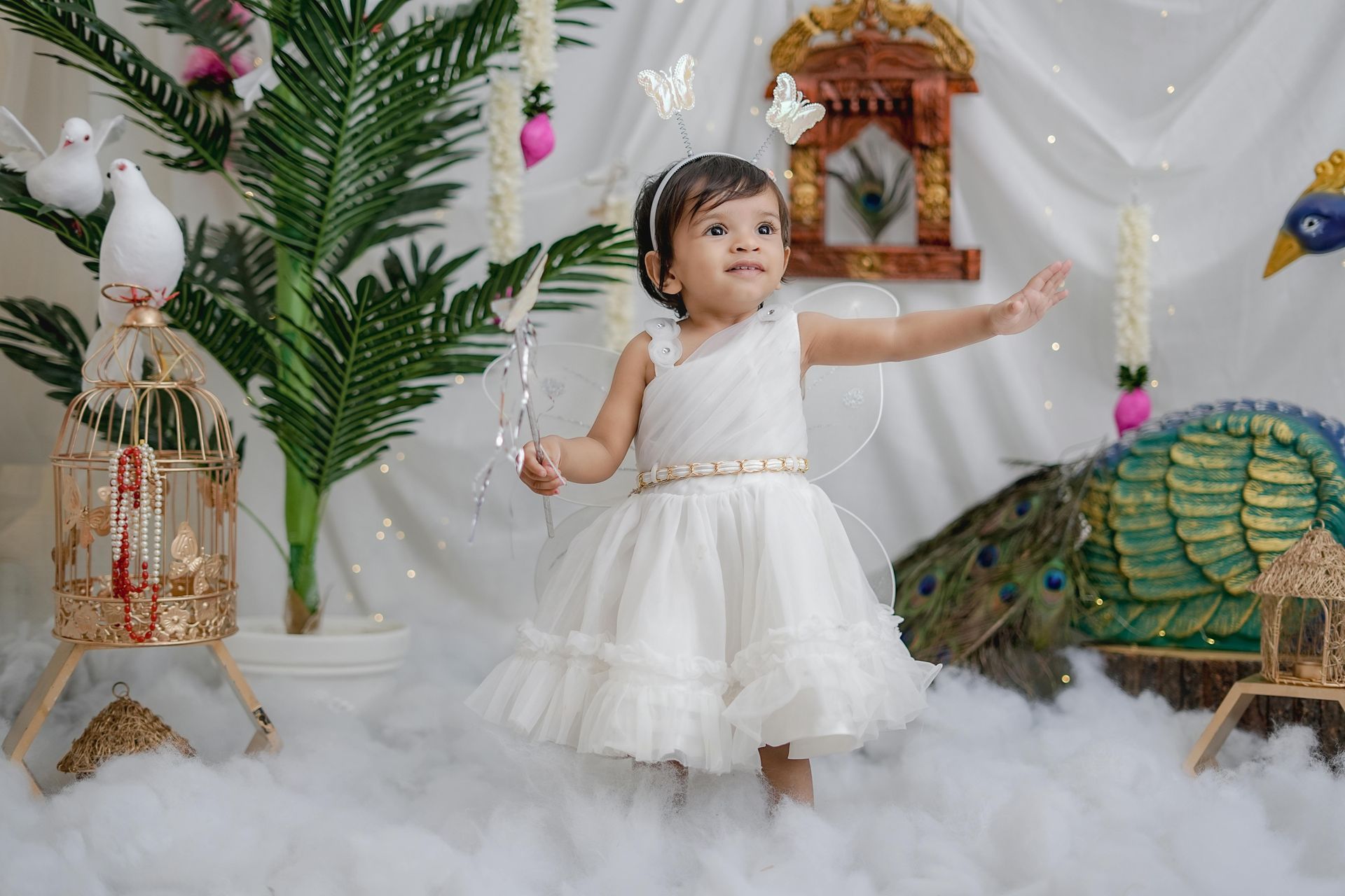 Child in a white dress standing in clouds, with decorative birds and greenery on a festive backdrop.