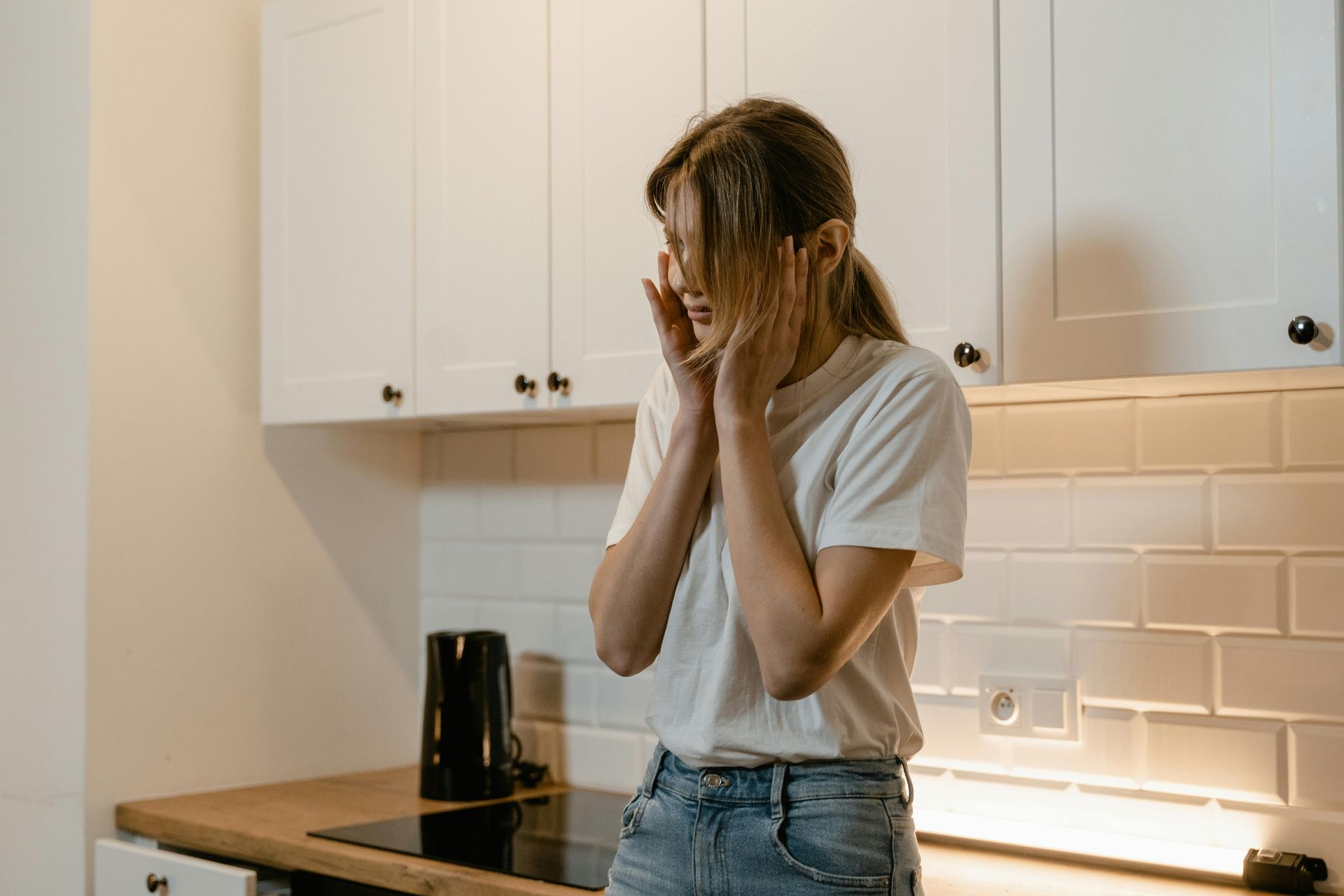 Person standing in a kitchen, covering their face with their hands.