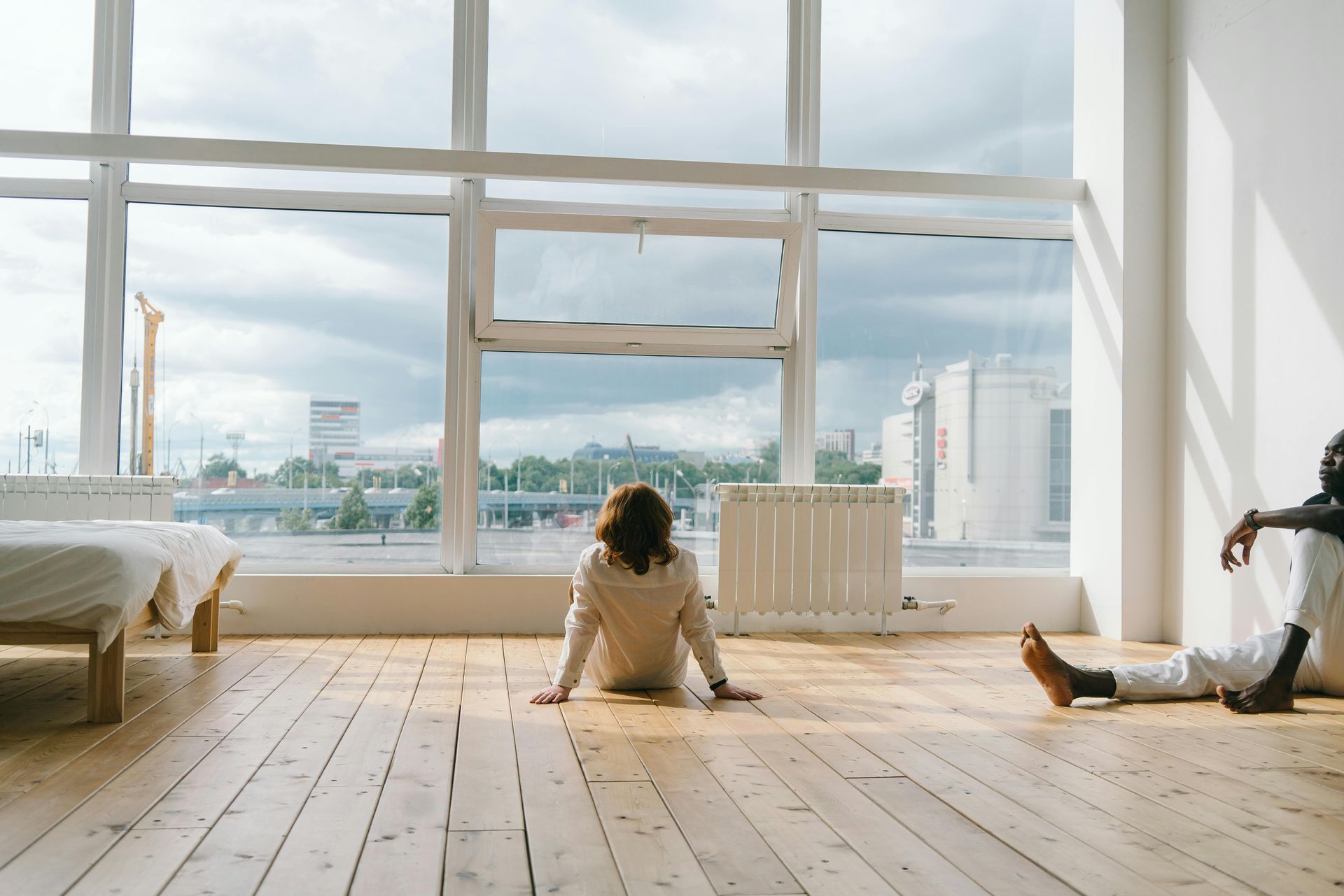 Bright room with large windows, two people sitting on a wooden floor overlooking a cityscape.