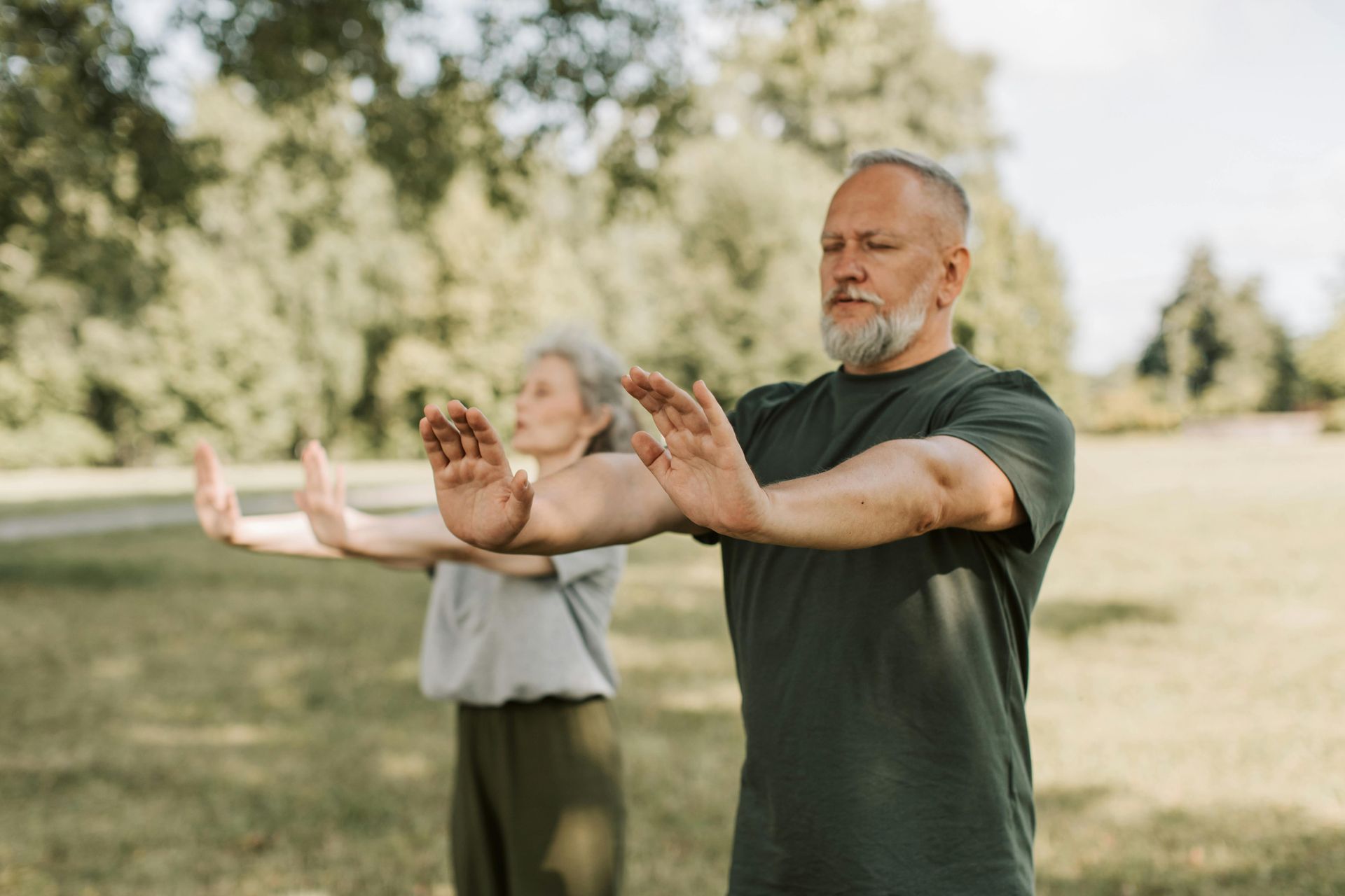 Two people practicing yoga outdoors in a grassy park, arms extended forward in a calm pose.