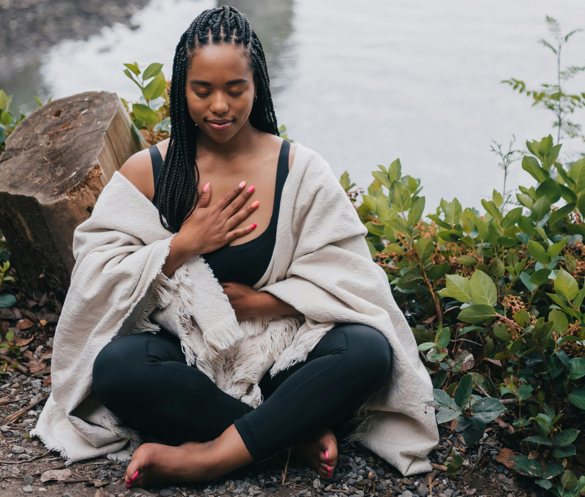 Woman meditating cross-legged outdoors beside rocks and greenery, hand on chest, wrapped in a light shawl