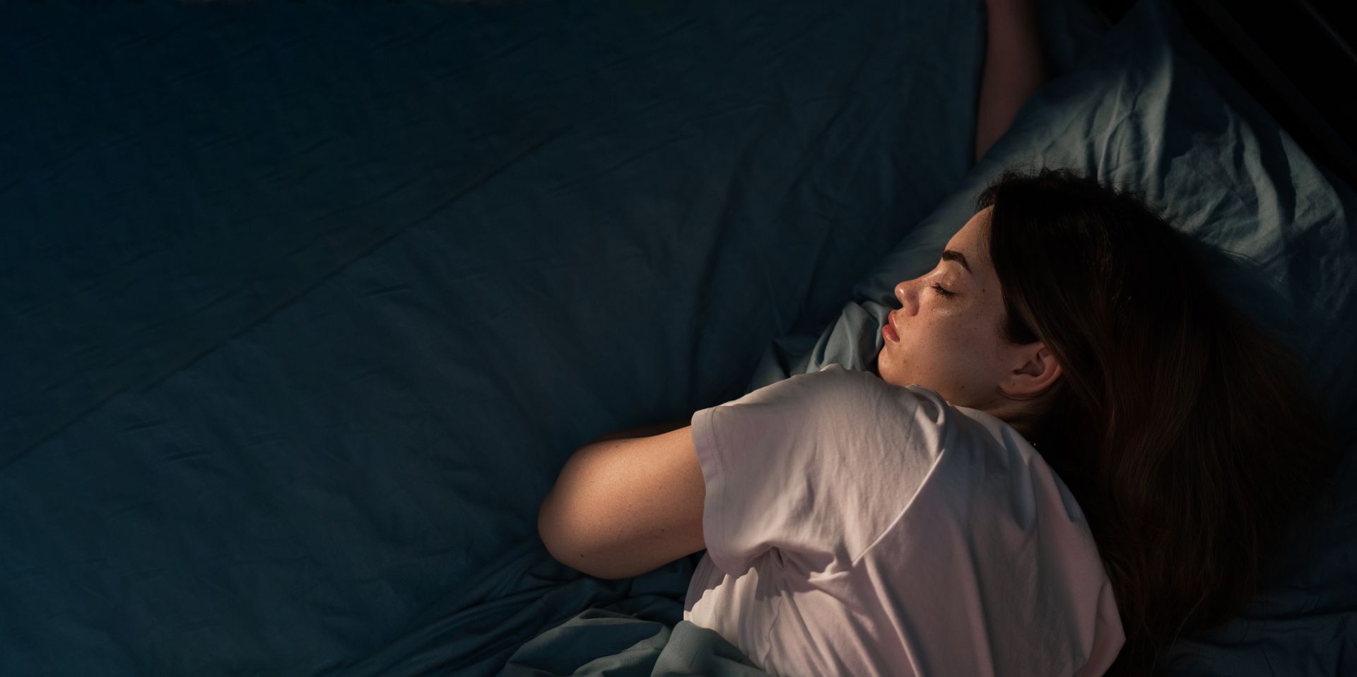 Person sleeping on a dark blue bed, wrapped in a white blanket, with soft light from the left.