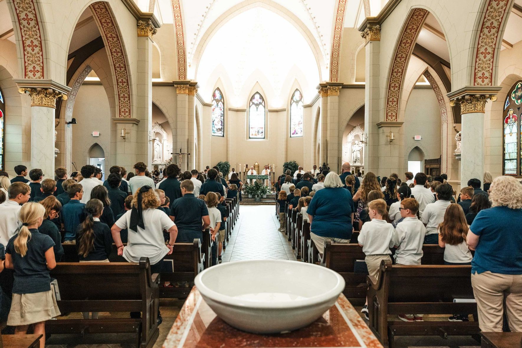 Congregation seated in a church, facing the altar, with a large baptismal font in the foreground.